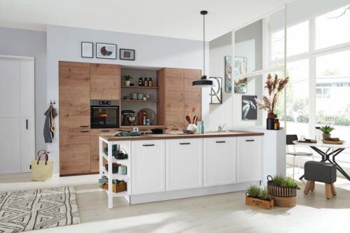 Die weiße Insel im Landhausstil ist ein toller Kontrast zur Schrankwand in Steineiche-Nachbildung. Um Harmonie zu schaffen, wird die gleiche Holz-Optik auch für die Insel-Arbeitsplatte verwendet. Schön wohnlich wirkt das offene Regal. Von Interliving. Foto: Interliving